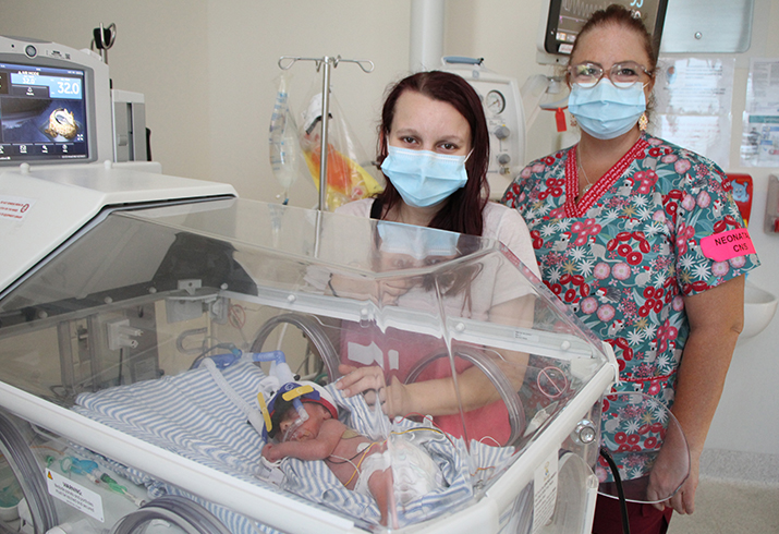 Premature baby lays in a neonatal cot. A young woman and a female nurse stand beside the cot.