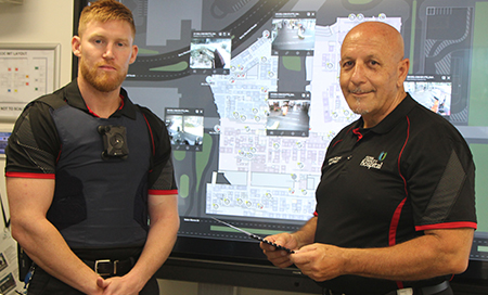 Two men stand in front of a large wall mounted screen which shows a map of a hospital campus.