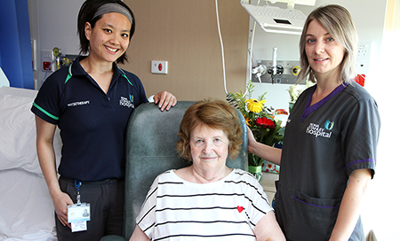 A young female physiotherapist and a young female nurse stand beside and older woman seated in a chair.