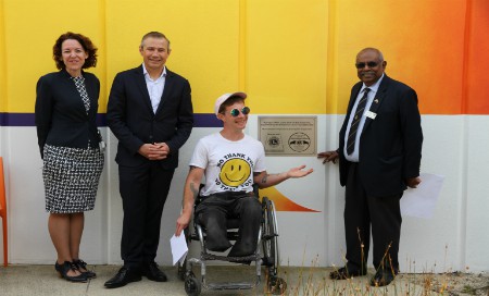 A woman and two men with a man in a wheelchair in front of a wall mural, pointing to a plaque