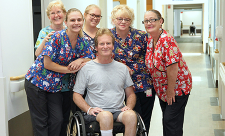 A young man sits in a wheelchair with five female nurses standing beside him. The nurses all wear bright surgical scrubs featuring Christmas designs.