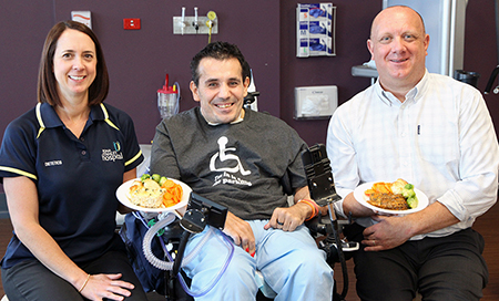 A man and woman holding plates of food sit next to a young man in a wheelchair